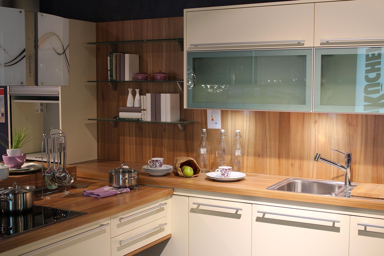 Cocina moderna con encimeras de madera, muebles claros, estantes de vidrio con vajilla y libros, fregadero de acero inoxidable y utensilios de cocina ordenados sobre la superficie.