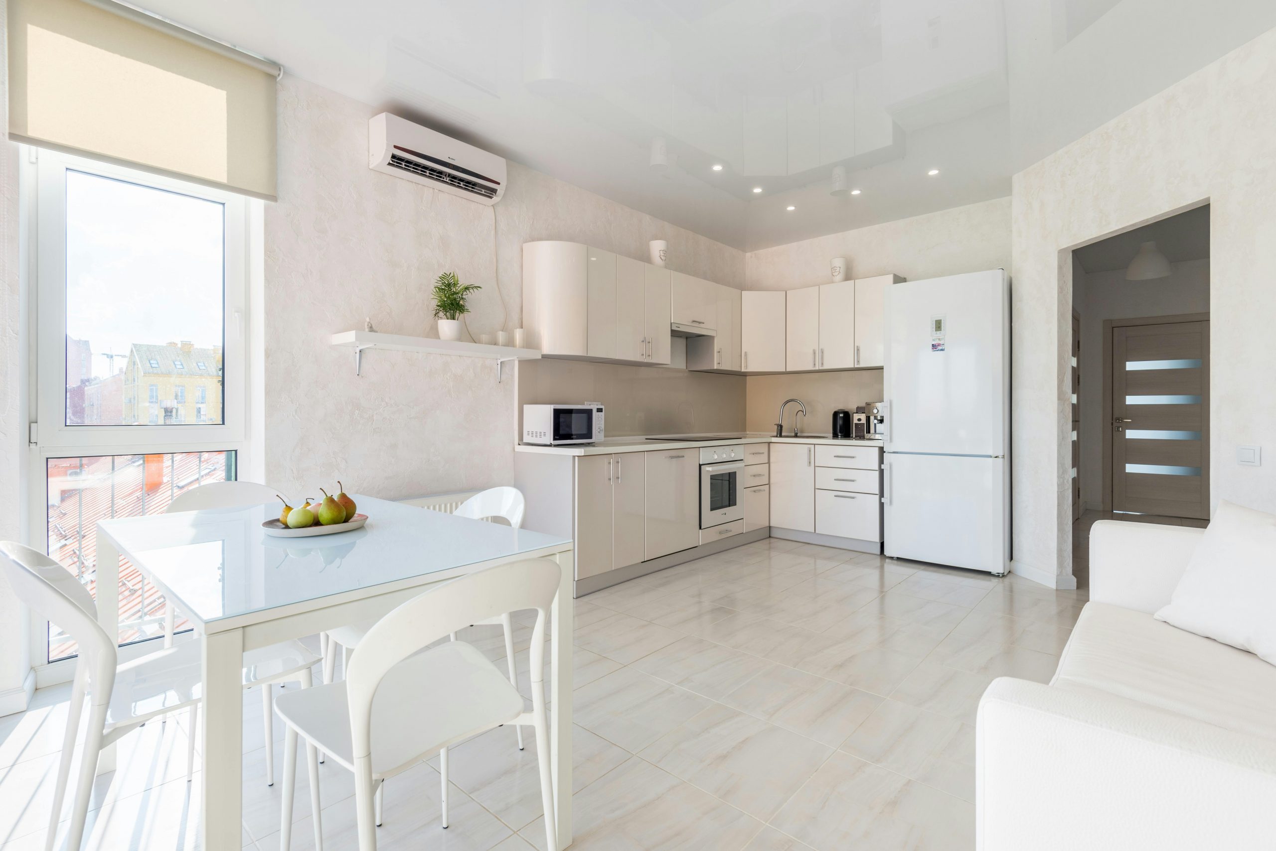 Cocina moderna y luminosa de estilo minimalista con mobiliario blanco, mesa de cristal con sillas a juego, electrodomésticos integrados, frigorífico grande y ventanal que aporta luz natural.