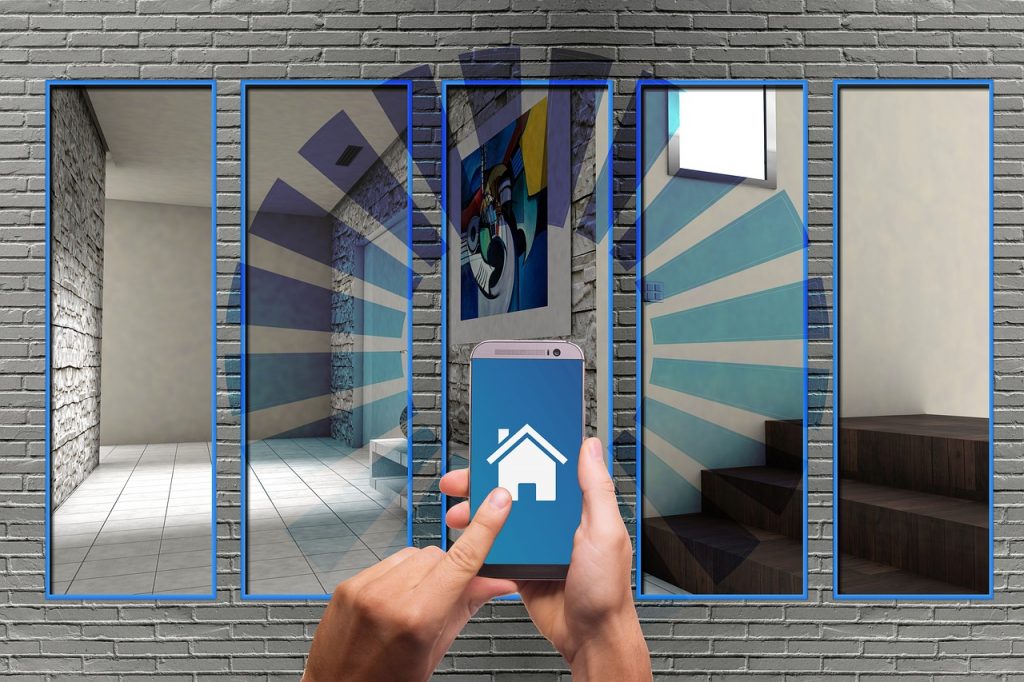 Manos sosteniendo un teléfono móvil con icono de casa en la pantalla, frente a un mural con imágenes de interiores, representando un concepto de hogar inteligente y control remoto de espacios.