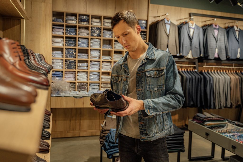Hombre joven con chaqueta vaquera observa un zapato en una tienda de moda masculina, rodeado de estanterías con calzado, camisas y trajes elegantes.