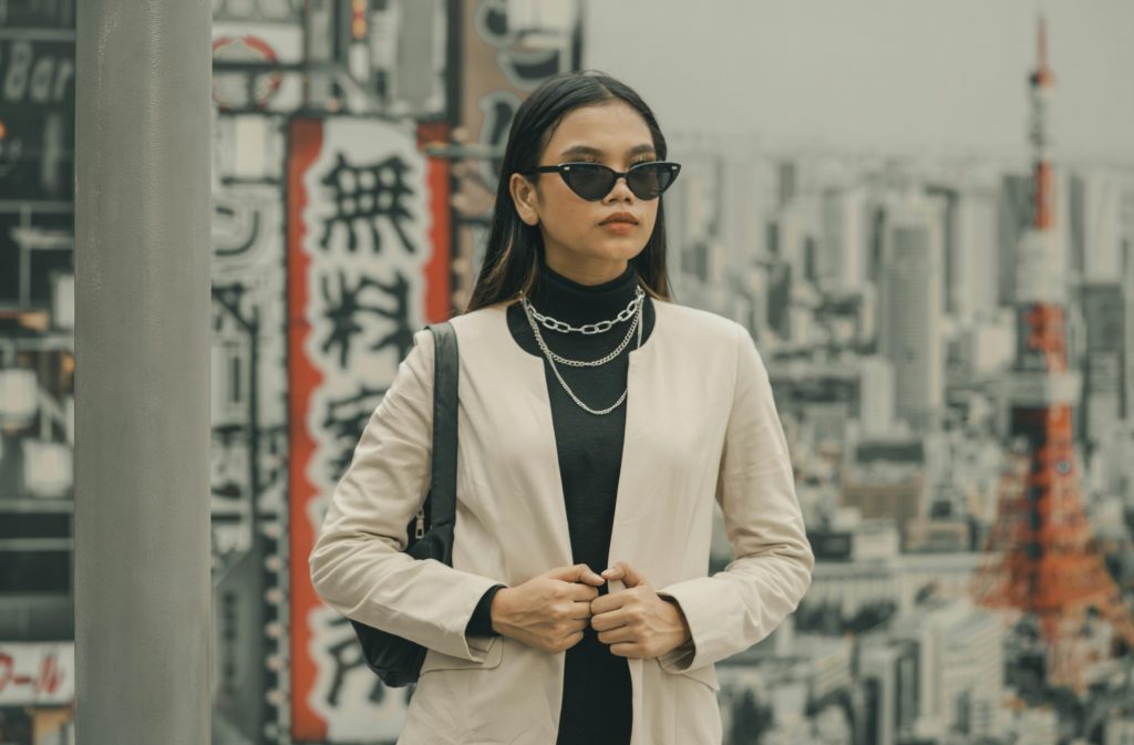 Mujer con gafas de sol negras y blazer claro posa con estilo en una ciudad asiática, con rascacielos y letreros japoneses de fondo.
