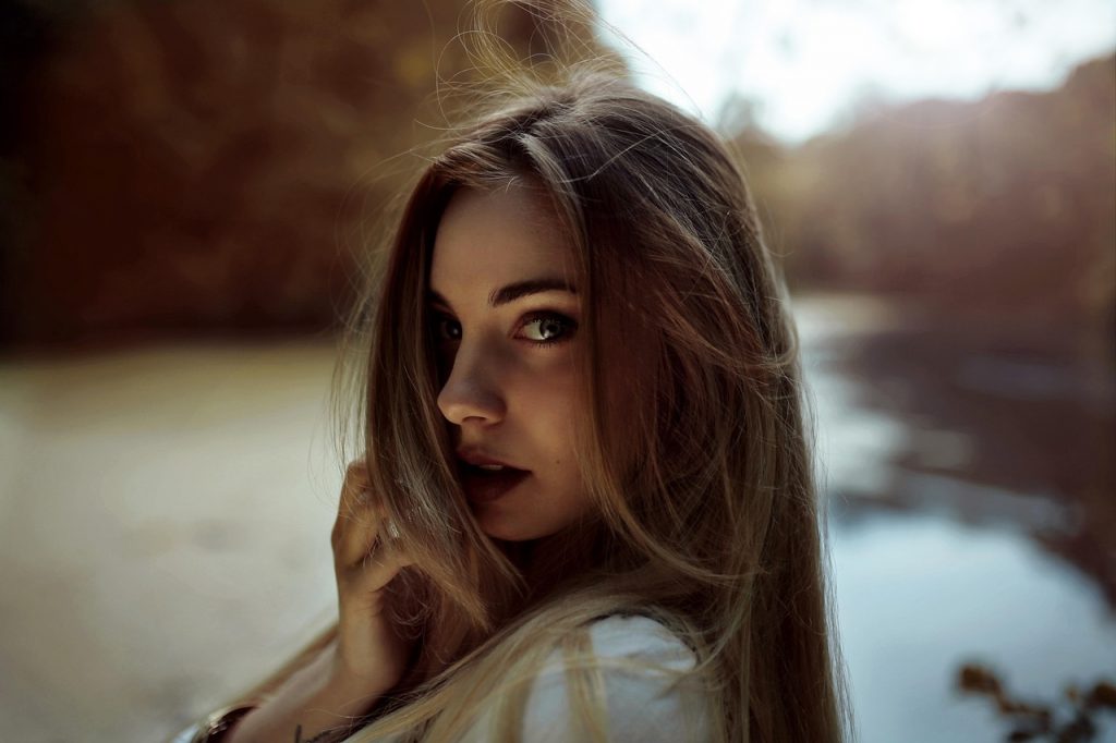 Mujer joven con cabello largo y suelto mira hacia la cámara en un entorno natural, con fondo desenfocado de agua y vegetación.