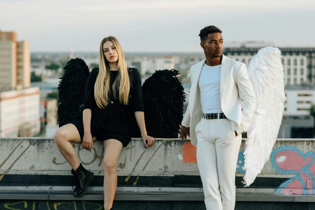 Dos jóvenes posan en una azotea con alas de ángel: la mujer viste de negro con alas negras y botas, mientras el hombre lleva un traje blanco con camiseta y alas blancas, ambos con un fondo urbano desenfocado.
