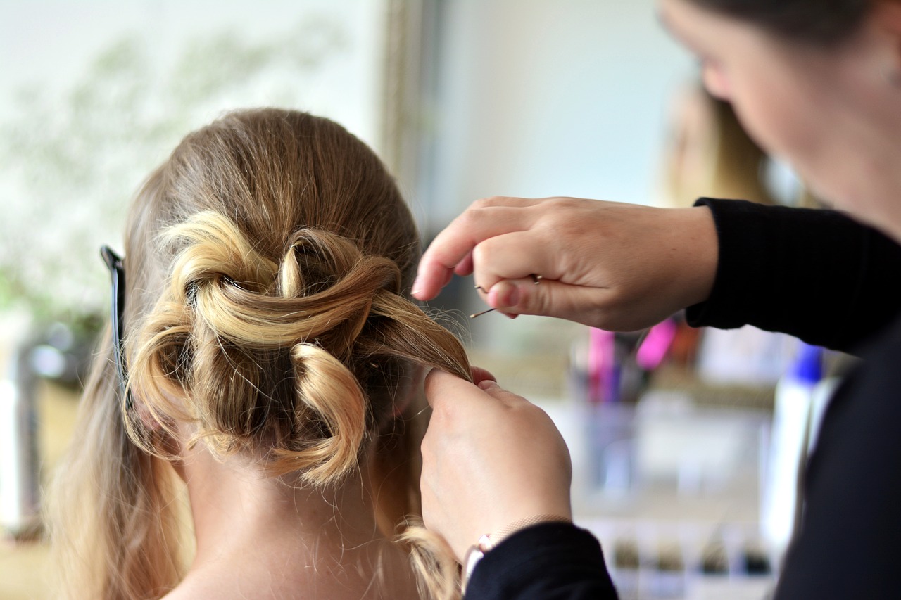 Manos de una estilista realizan un peinado recogido con rizos en el cabello rubio de una mujer, utilizando horquillas para fijar el estilo.