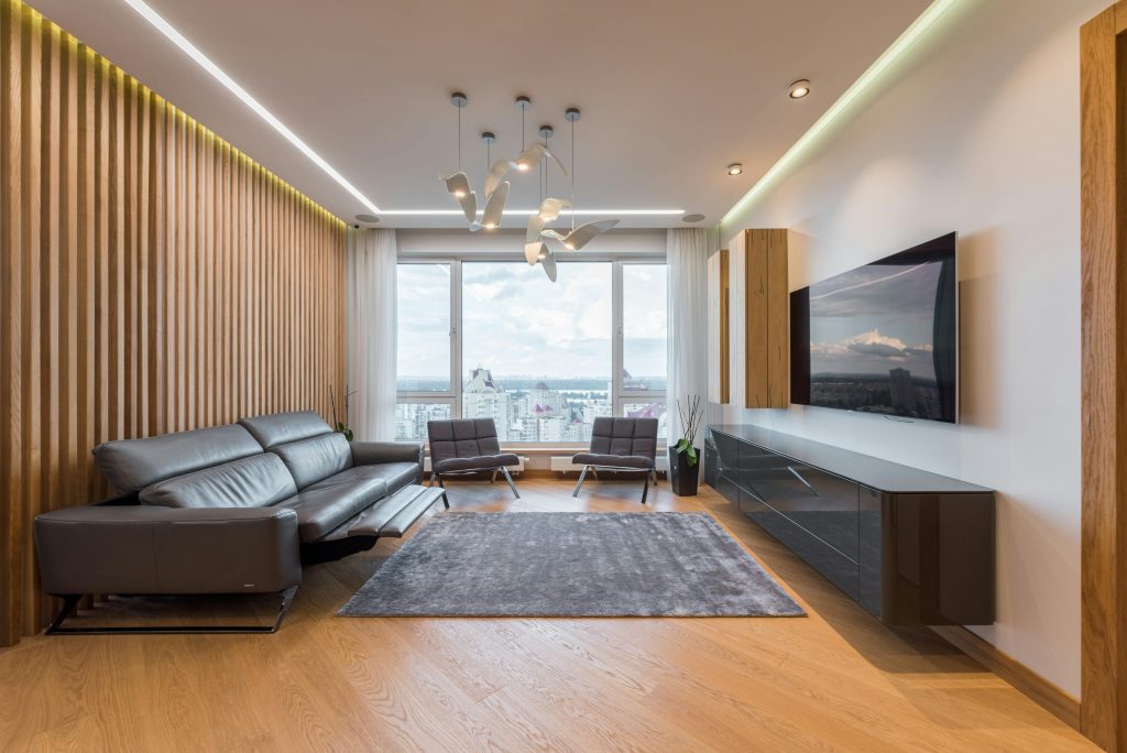 Salón moderno y luminoso con sofá de piel gris, alfombra gris sobre suelo de madera, gran ventanal con vistas a la ciudad, televisión de pantalla plana en la pared y decoración minimalista con detalles en madera.
