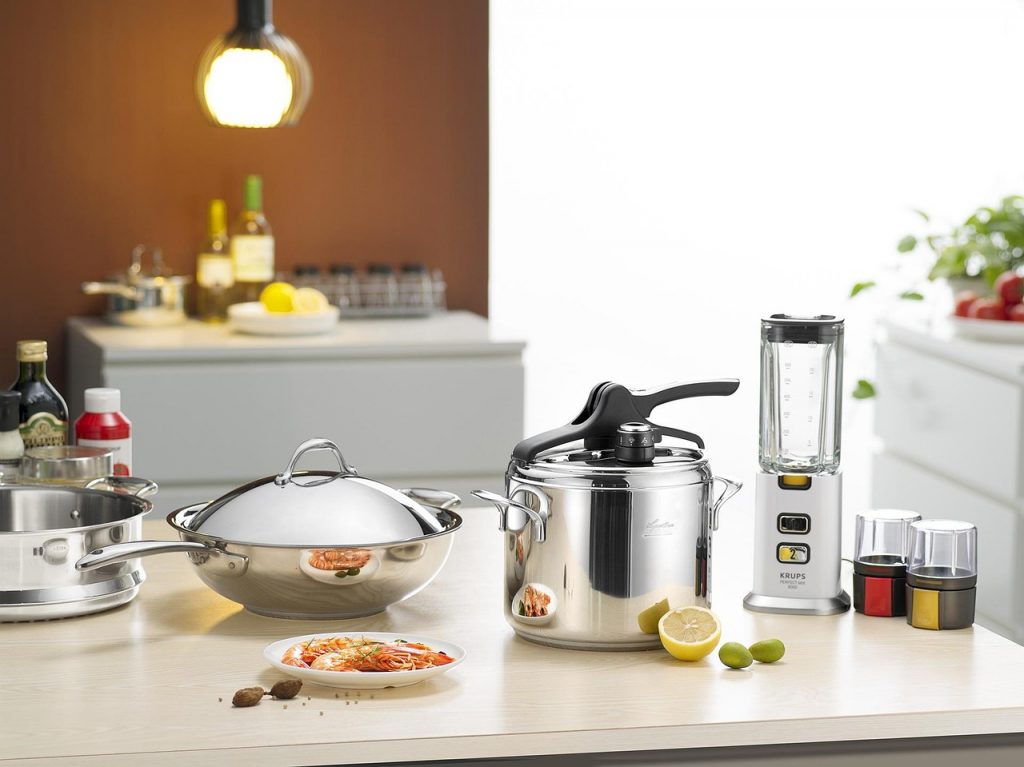Encimera de cocina con ollas de acero inoxidable, una licuadora eléctrica, especieros, medio limón, limas y un plato con mariscos preparados, bajo una lámpara colgante de luz cálida.