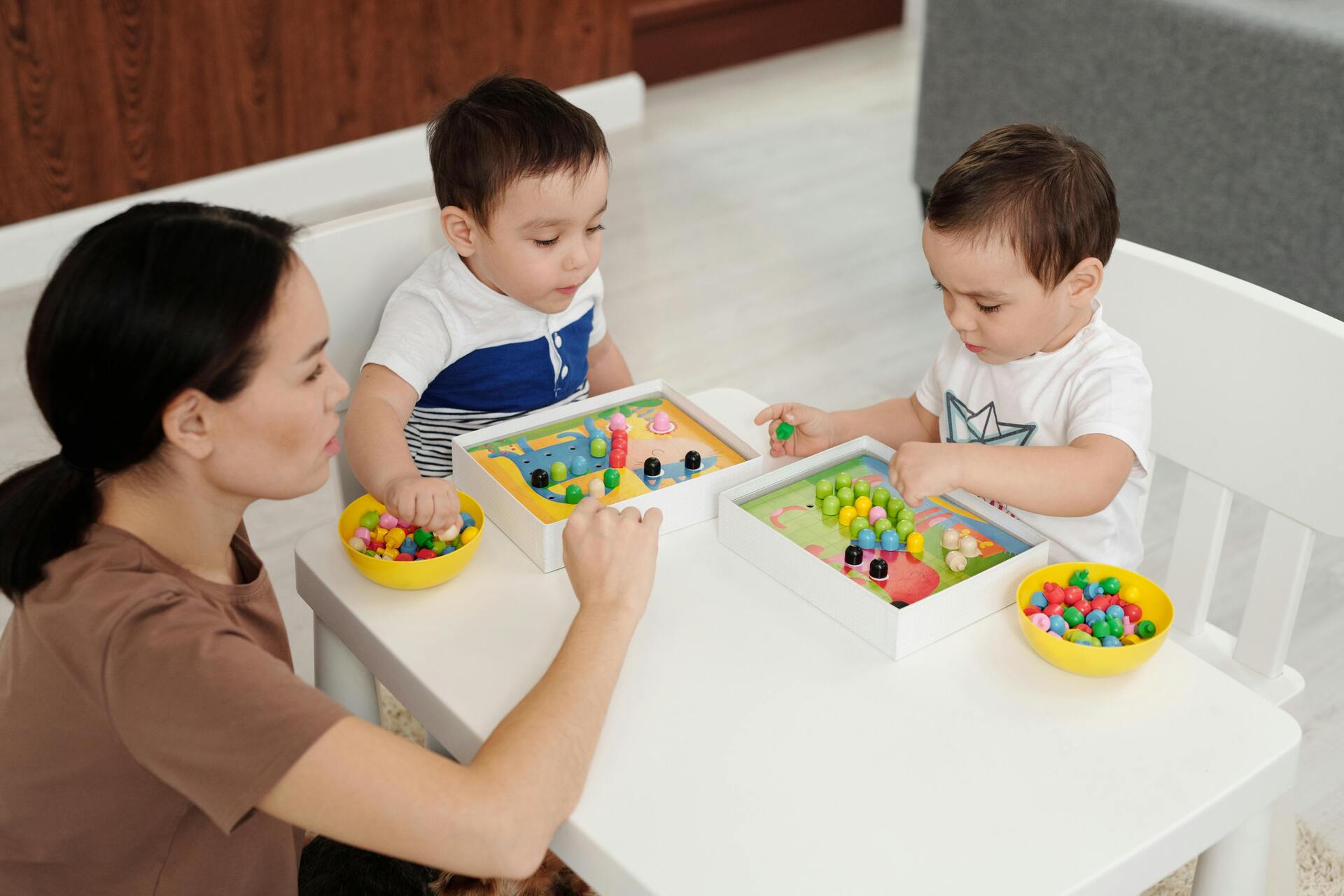 Dos niños pequeños jugando con juguetes educativos de cuentas de colores junto a una mujer en una mesa.